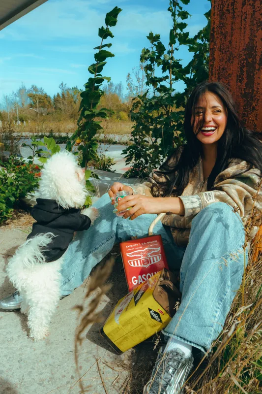 A smiling person sitting beside a white dog with Gasoline packaging at a vintage gas station.