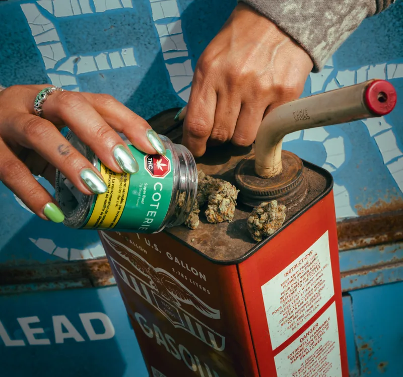 Hands tipping a green Coterie jar onto a vintage red Gasoline tin against a blue sign.