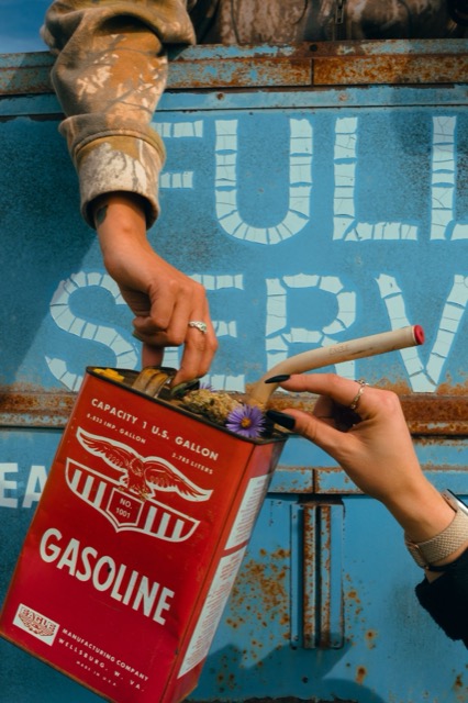 Hands holding a red Gasoline tin topped with cannabis flower and a purple flower against a blue sign.