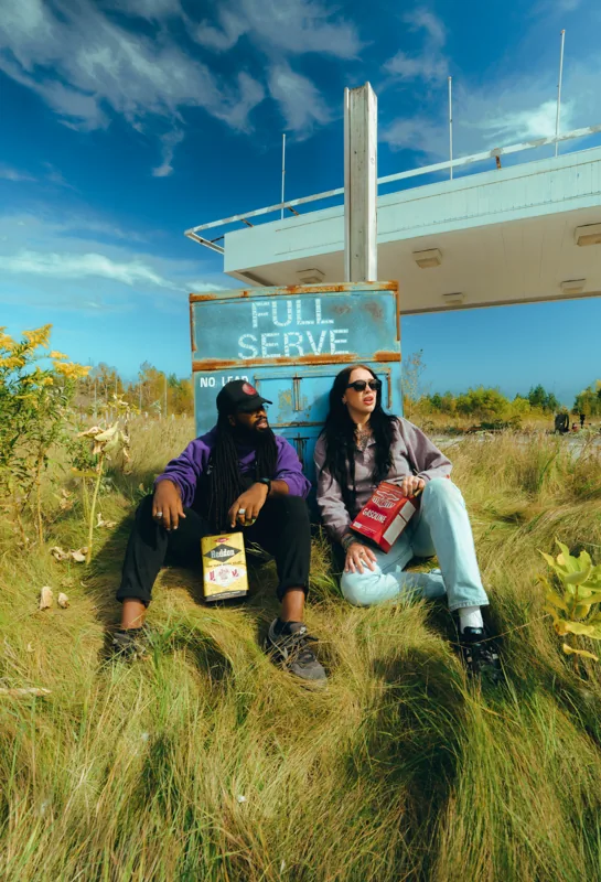 Two friends sitting in the grass beside a Full Serve sign at a vintage gas station.