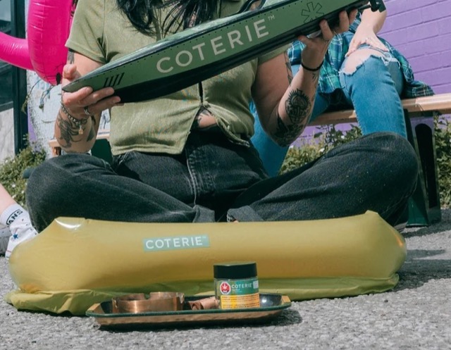 Hands holding a green Coterie prop above a yellow inflatable seat and product jar.