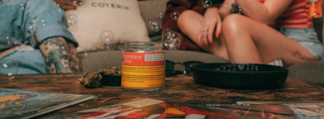A Coterie flower jar, cannabis flower and an ashtray on a coffee table in front of a sofa.